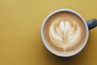 Creamy latte with intricate leaf pattern foam art in white ceramic cup against vibrant yellow background, top view composition for cafe and coffee shop marketing.