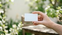 Mature male hand holding blank white business card outdoors in garden setting with blurred green foliage background for branding and marketing purposes.