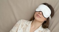 Young Caucasian woman relaxing with white sleep mask in bed wearing lace pajamas for rest and wellness routine.