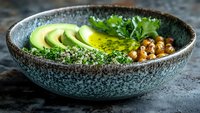 Healthy vegan bowl with sliced avocado, chickpeas, quinoa, and fresh herbs in rustic ceramic dish on dark stone background.