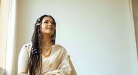 Young woman with braided hair and gold chain necklace wearing cream-colored shirt, looking upward thoughtfully against neutral background. Portrait for lifestyle and wellness content.