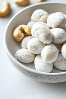 Mexican wedding cookies dusted with powdered sugar in speckled ceramic bowl, with cashews scattered nearby on white surface, macro food photography.