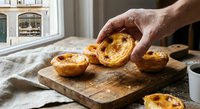 Hand picking golden Portuguese custard tart from rustic wooden board in cozy kitchen setting with warm natural lighting for bakery and culinary content.