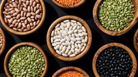 Assorted legumes and beans in wooden bowls including white beans, black beans, green lentils, and orange lentils arranged on dark background for cooking.
