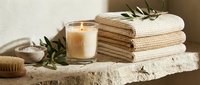 Cozy spa still life with lit candle, stacked waffle towels, olive branch, bath salt, and wooden brush on a stone shelf.
