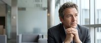 Thoughtful middle-aged Caucasian businessman in suit contemplating strategy in modern office environment with natural lighting and glass windows.