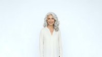 Mature woman with long silver wavy hair and fair complexion wearing cream button-up blouse, smiling at camera against white background for portrait and lifestyle use.