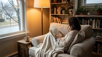 Young woman reading book on comfortable armchair in cozy home library corner with warm lamp light, bookshelves and window view creating peaceful atmosphere.