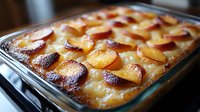 Homemade peach cobbler dessert in glass baking dish with golden brown crust and caramelized fruit slices arranged in rows, fresh from oven.