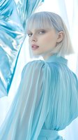 Young Caucasian woman with platinum blonde bob haircut in pleated turquoise dress poses against ethereal blue and white background for fashion and beauty content.