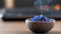 Burning blue resin in ceramic bowl with rising smoke, mystical aromatherapy setup on wooden surface against blurred laptop background for meditation practice.