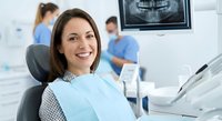 Happy female patient smiling in dental chair during checkup with dentist and assistant working in modern clinic background with x-ray monitor visible.