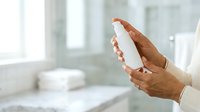 Mature woman with medium skin tone holding white cosmetic bottle in bathroom setting, displaying skincare product for beauty and wellness marketing purposes.