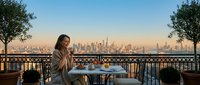 Young woman enjoying morning coffee on elegant balcony terrace overlooking city skyline at sunrise. Urban lifestyle and relaxation concept for hospitality design.