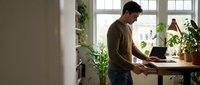 Young Caucasian man working on laptop at wooden desk in bright modern home office with plants creating productive workspace atmosphere for remote work.