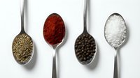 Four metal spoons filled with different spices and seasonings including herbs, paprika, black peppercorns and coarse salt on white background.