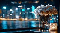 White roses in crystal vase against blurred city lights and water reflections at night, creating romantic atmosphere with turquoise bokeh background.