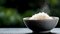 Steaming hot white rice in dark ceramic bowl on wooden table with blurred green nature background for healthy Asian cuisine and cooking concepts.