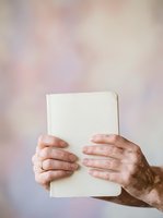 Mature adult hands holding blank white notebook against soft blurred background, suitable for education, publishing, writing, and business concepts.