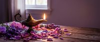 Antique brass oil lamp with glowing flame surrounded by purple flower petals on rustic wooden table near window creating mystical Arabian nights atmosphere.