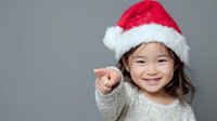 Smiling Asian girl in Santa hat pointing finger at viewer against gray background, wearing cozy sweater for holiday season celebration.