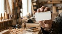 Craftsman holding blank white card in woodworking workshop with tools and wood materials, suitable for business messaging and trade promotion.