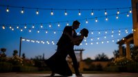 Romantic couple dancing under string lights at twilight outdoor wedding celebration with warm glowing bulbs creating magical atmosphere for special occasion.