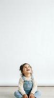 Toddler girl with dark curly hair, South Asian ethnicity, wearing denim overalls and cream ribbed long-sleeve shirt, sitting on wooden floor against white wall, looking upward with wonder and joy.