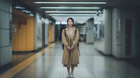 Young Asian woman in beige trench coat standing in modern subway station corridor with fluorescent lighting and tiled floors for urban lifestyle concepts.