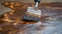 Wide paintbrush with silver metal ferrule applying protective varnish coating on wooden surface in warm brown tones, close-up macro shot showing brush bristles detail.