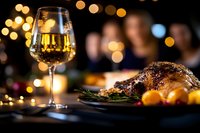 Glass of white wine and roasted duck with citrus garnish on dark table against blurred festive lights background for restaurant menu and holiday dining.
