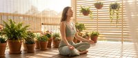 Young woman practicing yoga meditation in lotus pose surrounded by potted plants on sunny wooden deck with warm natural lighting and hanging planters.