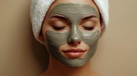 Young woman with clay facial mask wearing white towel on head during spa treatment, eyes closed with serene expression on beige background.