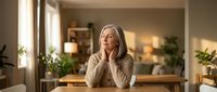 Thoughtful mature blonde woman sitting at wooden table in bright modern home interior with natural sunlight and green plants.