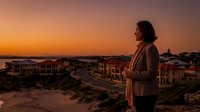 Woman in casual blazer standing on coastal cliff overlooking residential neighborhood at golden hour sunset, contemplative pose for lifestyle and travel concepts.