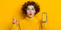 Surprised young woman with curly hair in yellow sweater showing smartphone screen with blank display against bright yellow background for advertising.