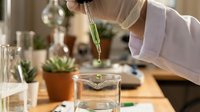 Scientist in white lab coat dropping green liquid into glass beaker during chemical experiment, conducting research with plants and botanical samples on wooden laboratory table.