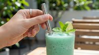 Cropped hand holding stainless steel straw in green smoothie drink with mint leaf garnish outdoors, sustainable beverage lifestyle concept for eco-friendly dining and reusable drinkware.