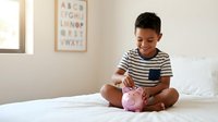 Young boy, approximately 6-8 years old, Hispanic ethnicity, sitting on white bed in bright bedroom, inserting coin into pink piggy bank, wearing striped shirt, learning financial literacy and saving money habits.