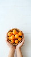Overhead view of hands holding woven basket filled with fresh oranges against white background, isolated for design use on merchandise and promotional materials.