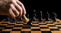 Male hand moving wooden chess piece on checkered board during strategic game play against black pieces on dark background.