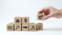 Hand placing wooden cube with gear icon onto stack of business symbol blocks representing strategy and growth concepts for corporate planning.
