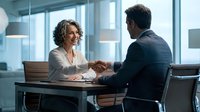 Professional businesswoman and businessman shaking hands across conference table in modern office meeting room with glass walls and city view.