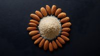 Whole almonds arranged in sunflower pattern around pile of ground almond flour on dark background, creative food photography composition for healthy eating concept.