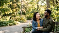 A diverse couple sits together on a park bench surrounded by lush green foliage, sharing a joyful moment outdoors. The woman, appearing to be of Asian descent, wears a denim jacket over a yellow top and smiles warmly while the man, appearing to be of African descent, wears an olive green sweater. Dappled sunlight filters through the trees, creating a warm, romantic atmosphere perfect for relationship, lifestyle, and outdoor leisure concepts.