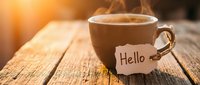 Steaming coffee cup with Hello tag on rustic wooden table in warm morning sunlight, cozy greeting concept.