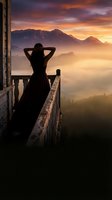 Woman in silhouette standing on balcony overlooking misty mountain landscape at sunrise, contemplative pose with hands behind head, dramatic golden sky and fog-covered valleys below.