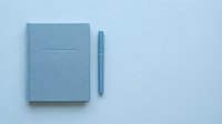 Closed blue notebook with pen on light blue background, flat lay composition for stationery, office supplies, and business organization concepts.