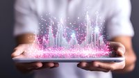 Futuristic holographic city skyline projection emerging from tablet device with glowing pink and blue particles held by hands against dark background.