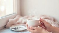 Cozy morning scene with hands holding white coffee cup in soft natural light by window with warm blanket creating peaceful atmosphere for relaxation and comfort.